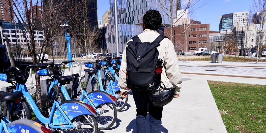 Nikhil with his back towards the camera walking alongside a rack of Blubikes on MIT Sloan's campus. In the background, the skyline of Kendall Square.