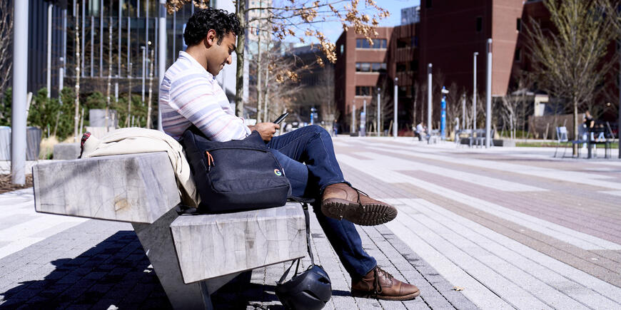 Nikhil sitting on an outdoor bench on MIT's campus. He is looking down at his phone, next to him is his backpack and helmet.