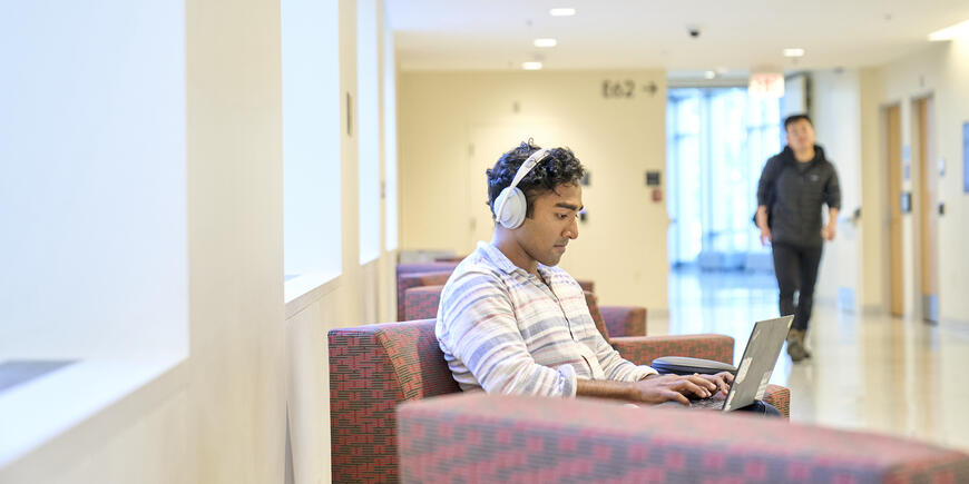 Nikhil sitting on an arm chair in a brightly lit hallway at MIT Sloan. He has headphones on and is looking down at his laptop. In the background, empty arm chairs and a person is walking down the hall. 