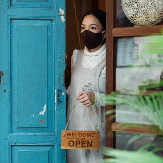 Woman in a face mask opening the door of a business holding a "Welcome, we're open" sign looking tentative and unsure.