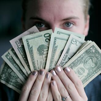 Image of the face of a woman who is holding U.S. currency fanned out in her hands, obscuring the lower half of her face. 