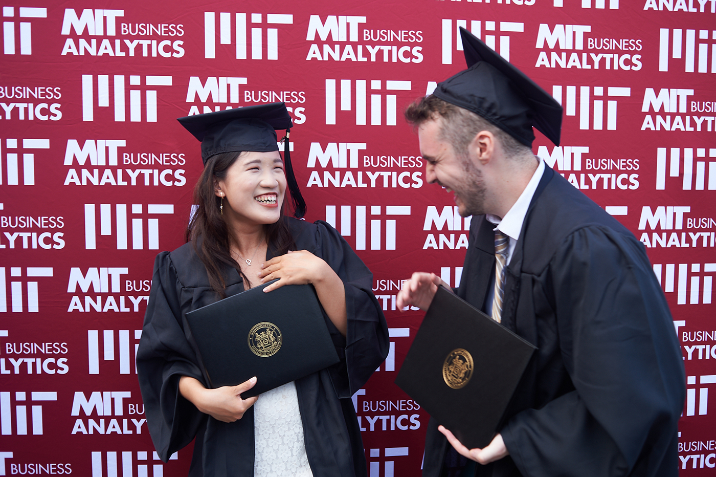 Man and woman in graduation cap and gown each holding their diplomas, facing each other and smiling. A red background with the MIT Master of Business Analytics in white lettering.