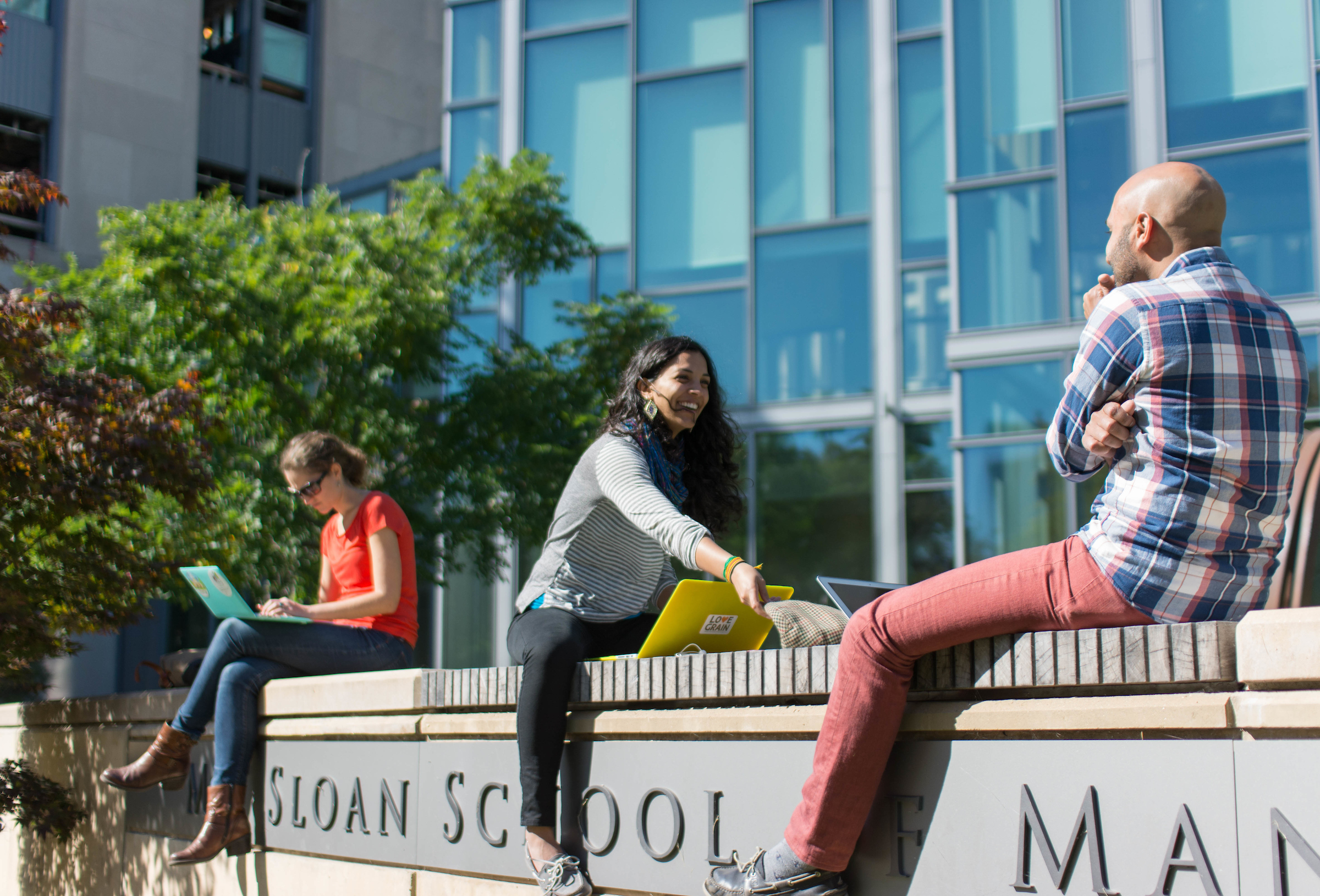 Three young adults sit on a wall outside the MIT Sloan School of Management