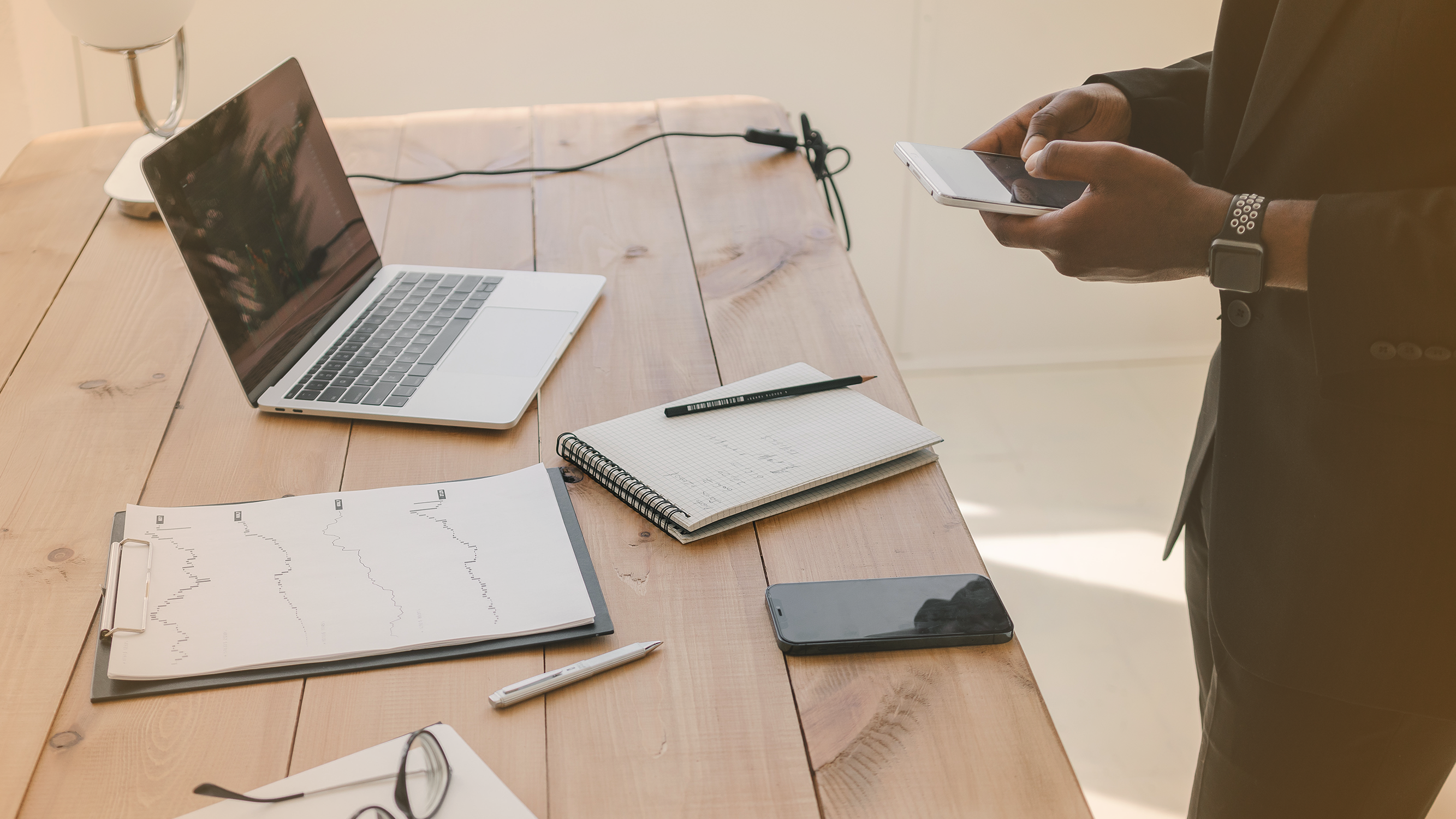 Person standing at desk with laptop and notebook looking at a phone