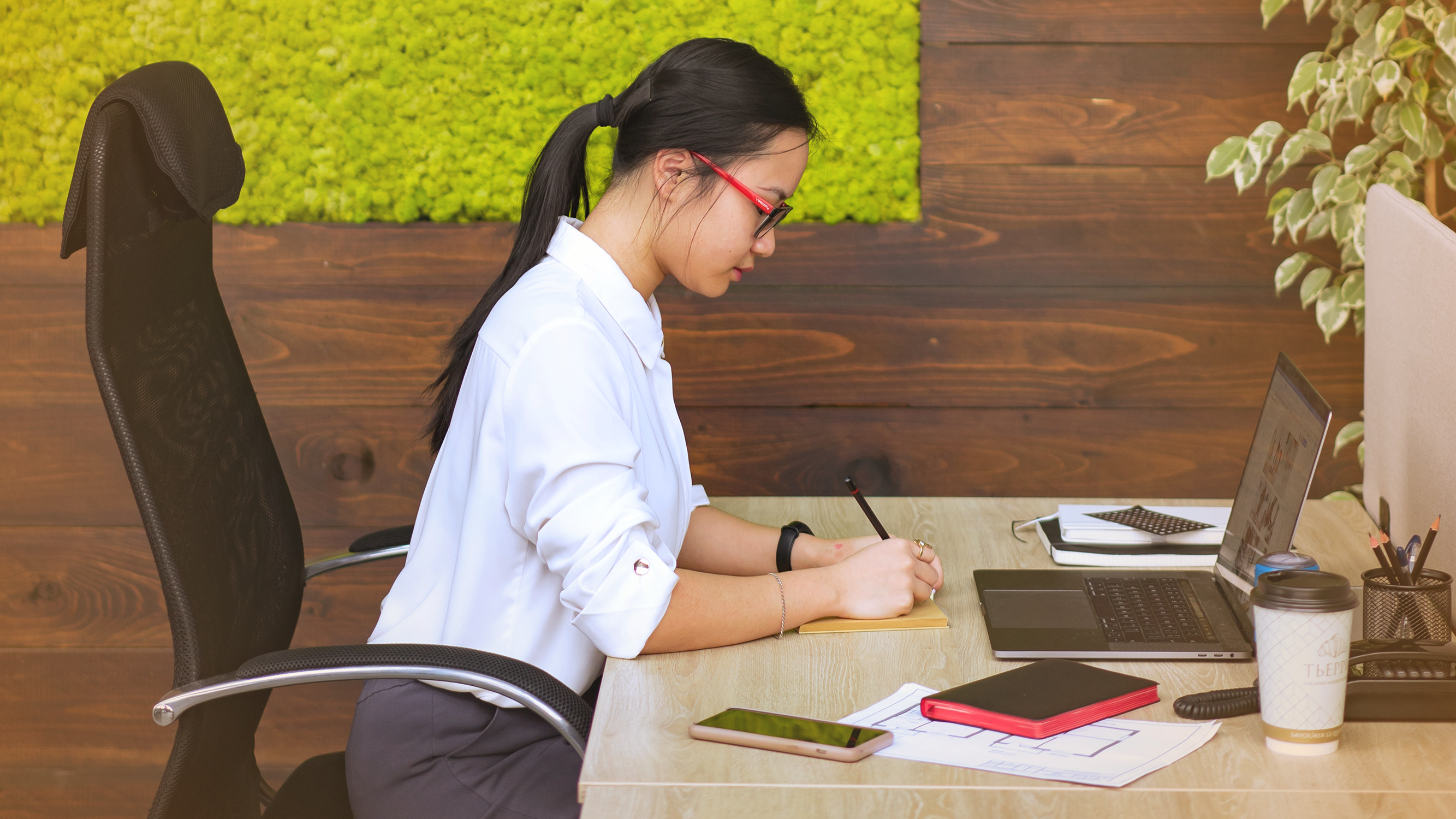 Woman working at a desk in an office with a laptop, pen and paper