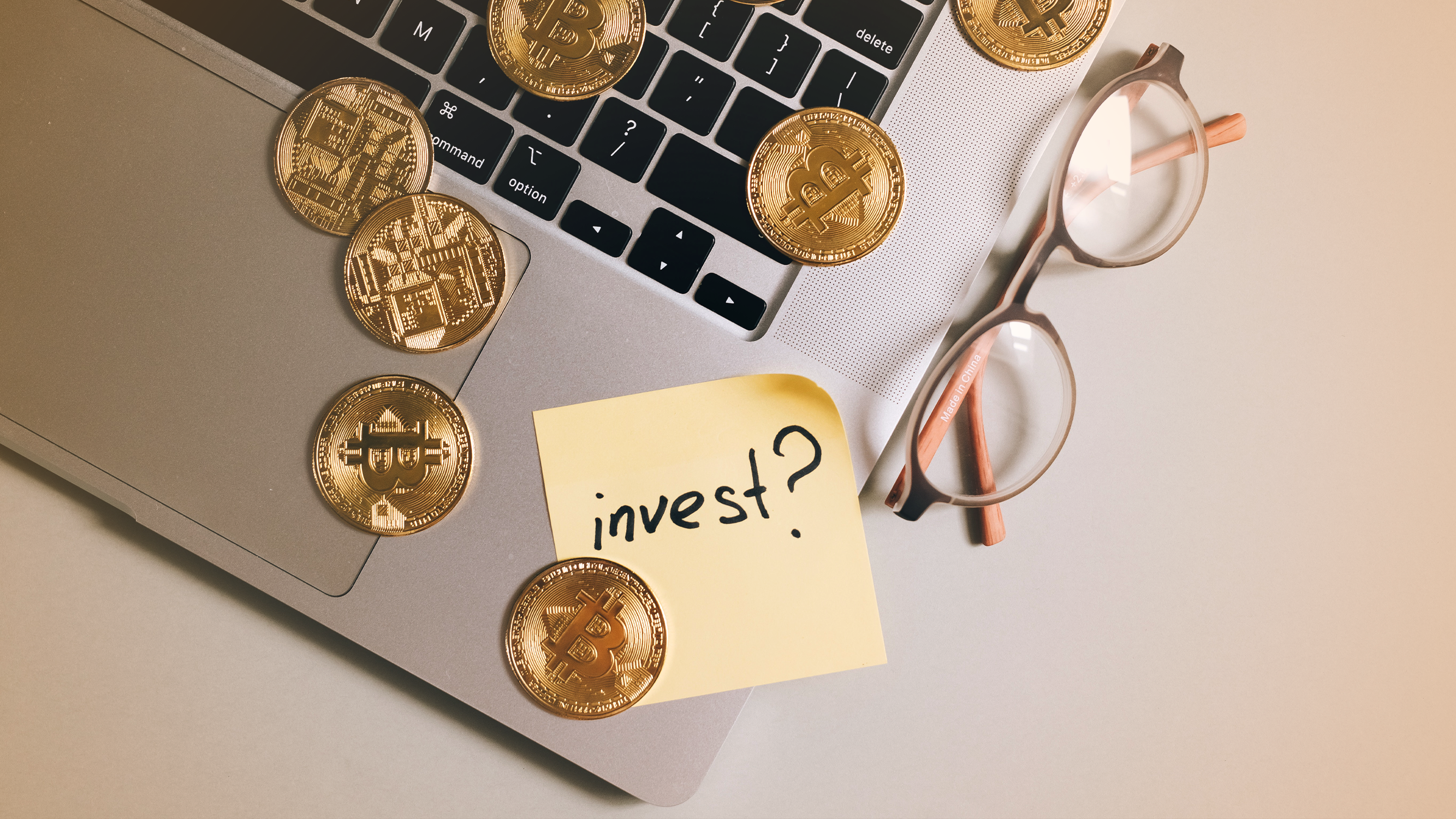 Popular cryptocurrency coins on a laptop keyboard with a sticky note reading "Invest?" Includes bitcoin, ethereum, solana