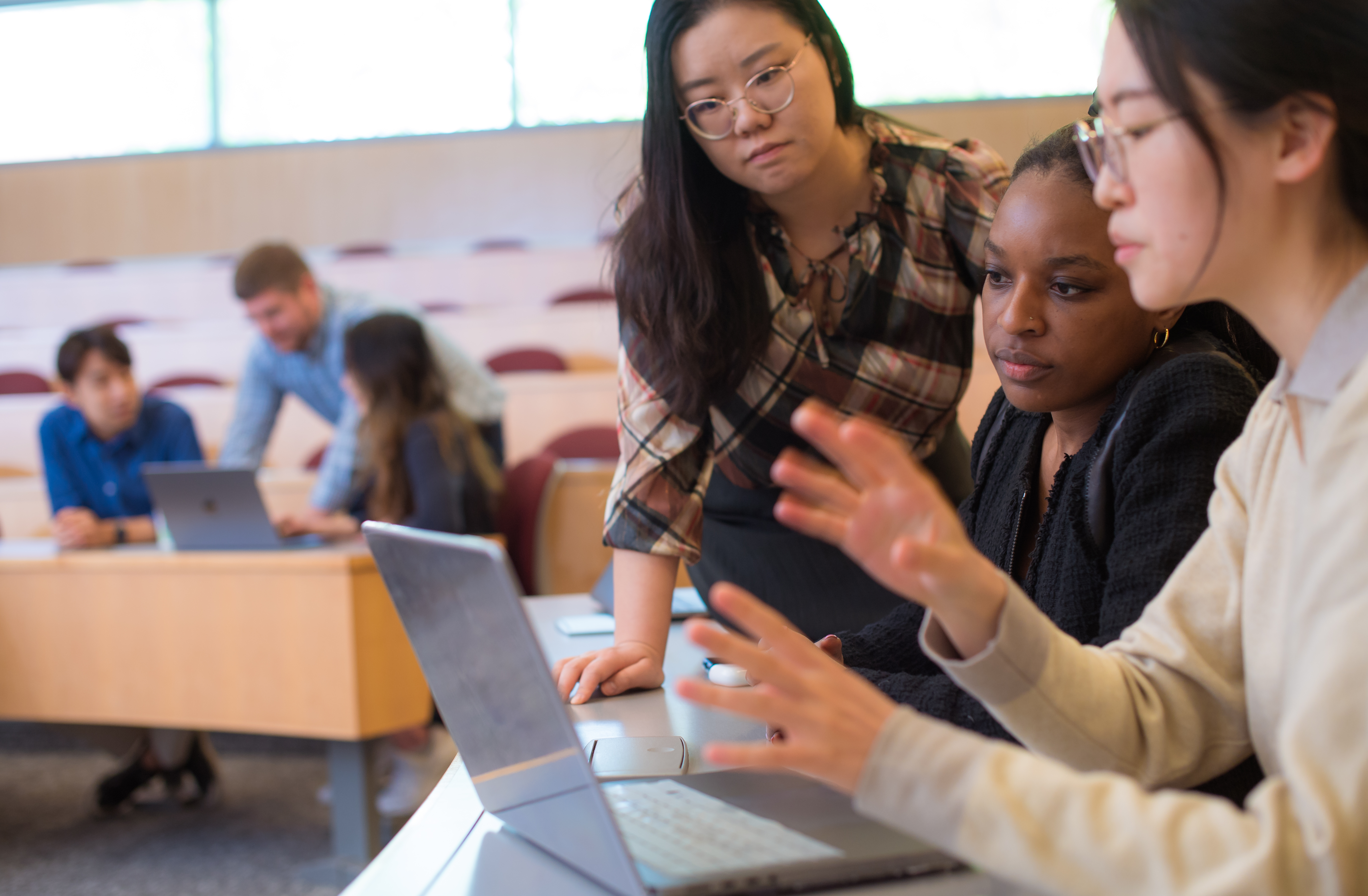 A group of three women looking at a laptop in a classroom and a group of three students in the background