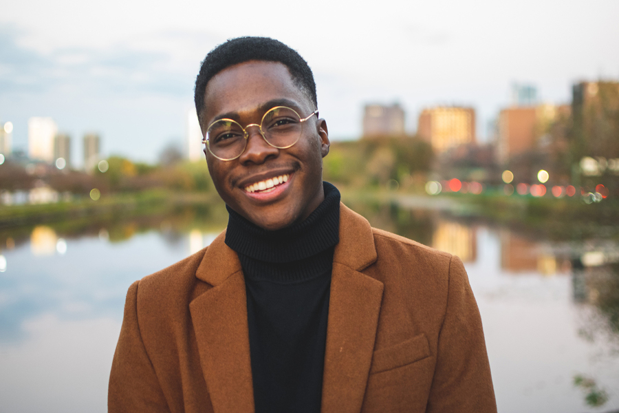 Man in glasses wearing suit poses in front of a river and cityscape