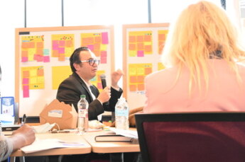 Two people seated, post it notes suggesting workshopping in background
