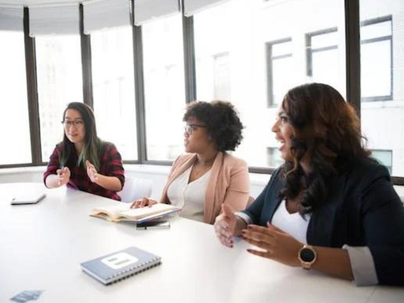 Three woman in discussion at white table in discussion room, windows behind them