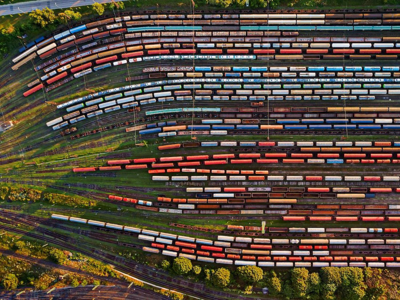 Many railroad tracks in rows from top to bottom with many multicolored cars on each, green landscape behind..