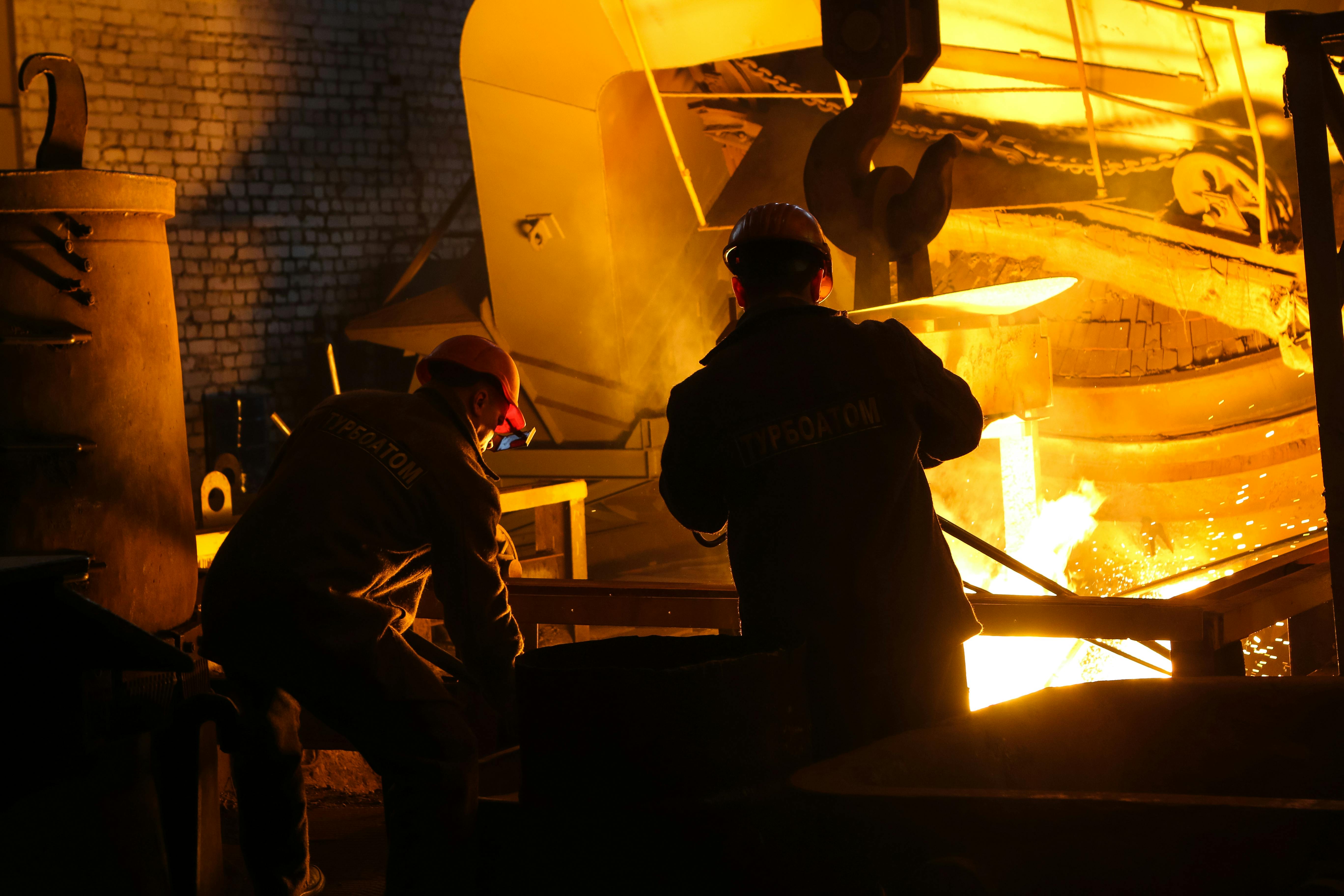 Image of two workers in helmets standing near a melt pool in steel manufacturing; what appears to be molten metal is visible in the background.