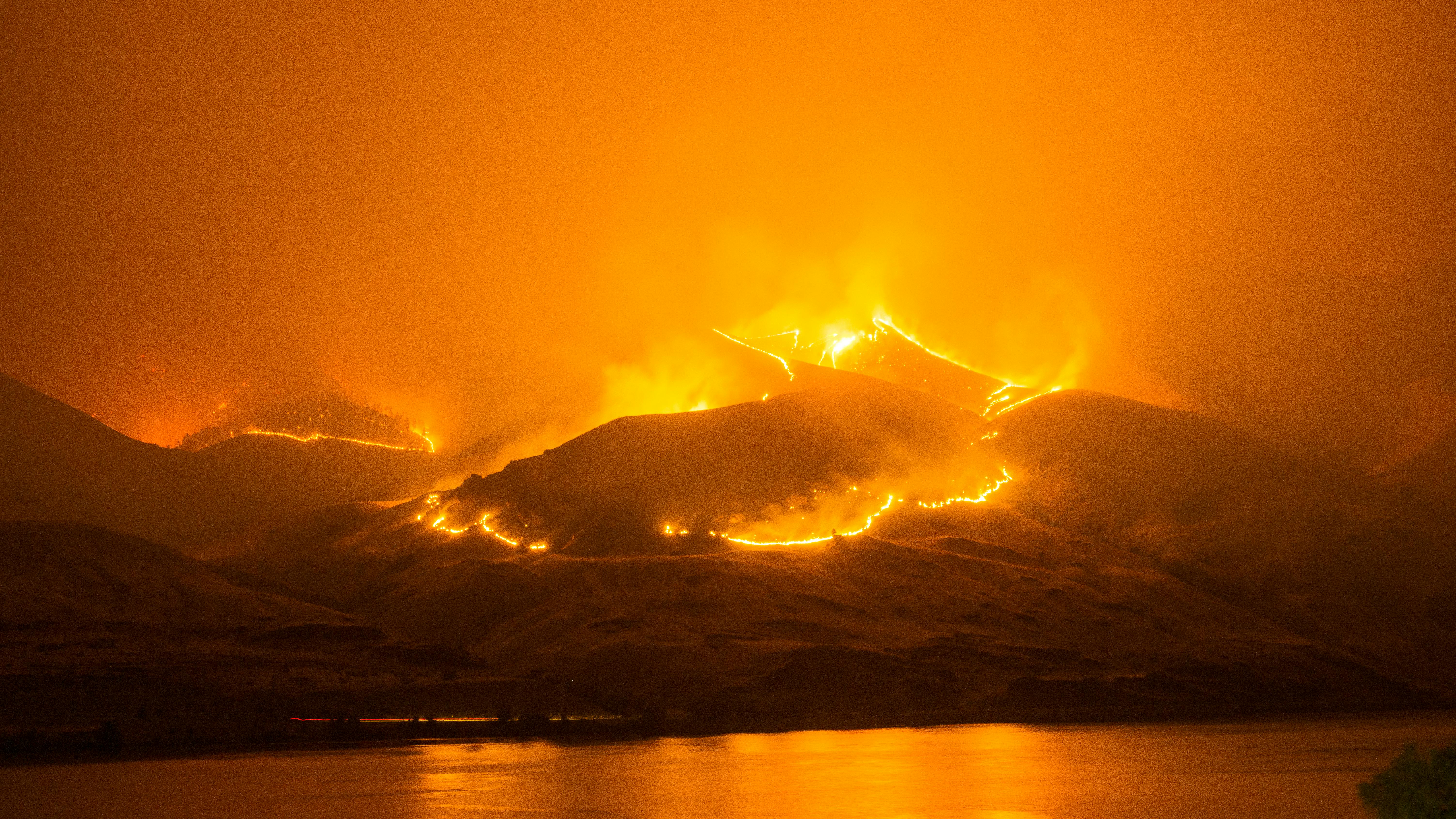 Image of a wildfire coursing across mountain tops at night, by a body of water.