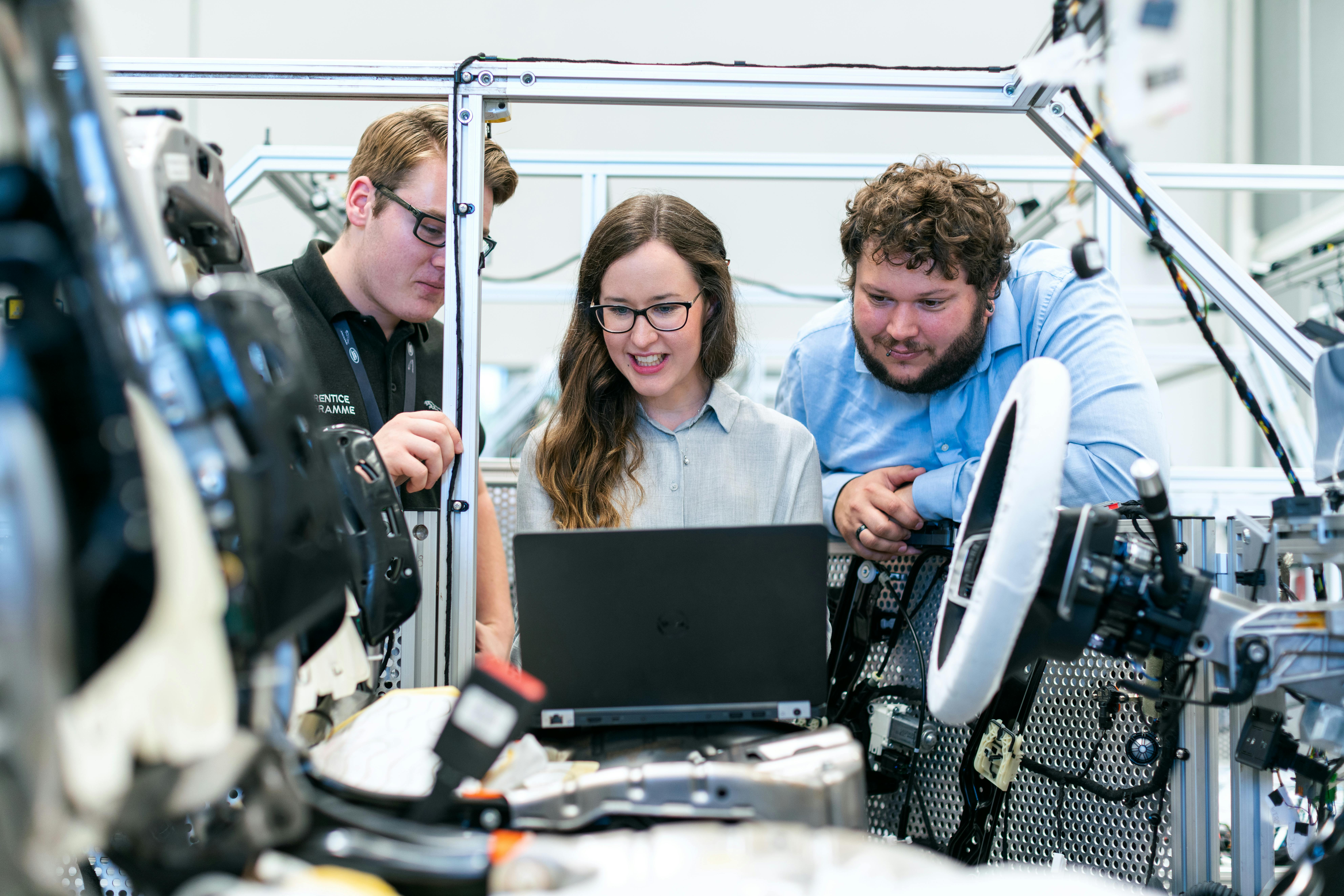 Group of three people, 2 men and 1 woman in the middle, looking at a laptop, surrounded by tech.