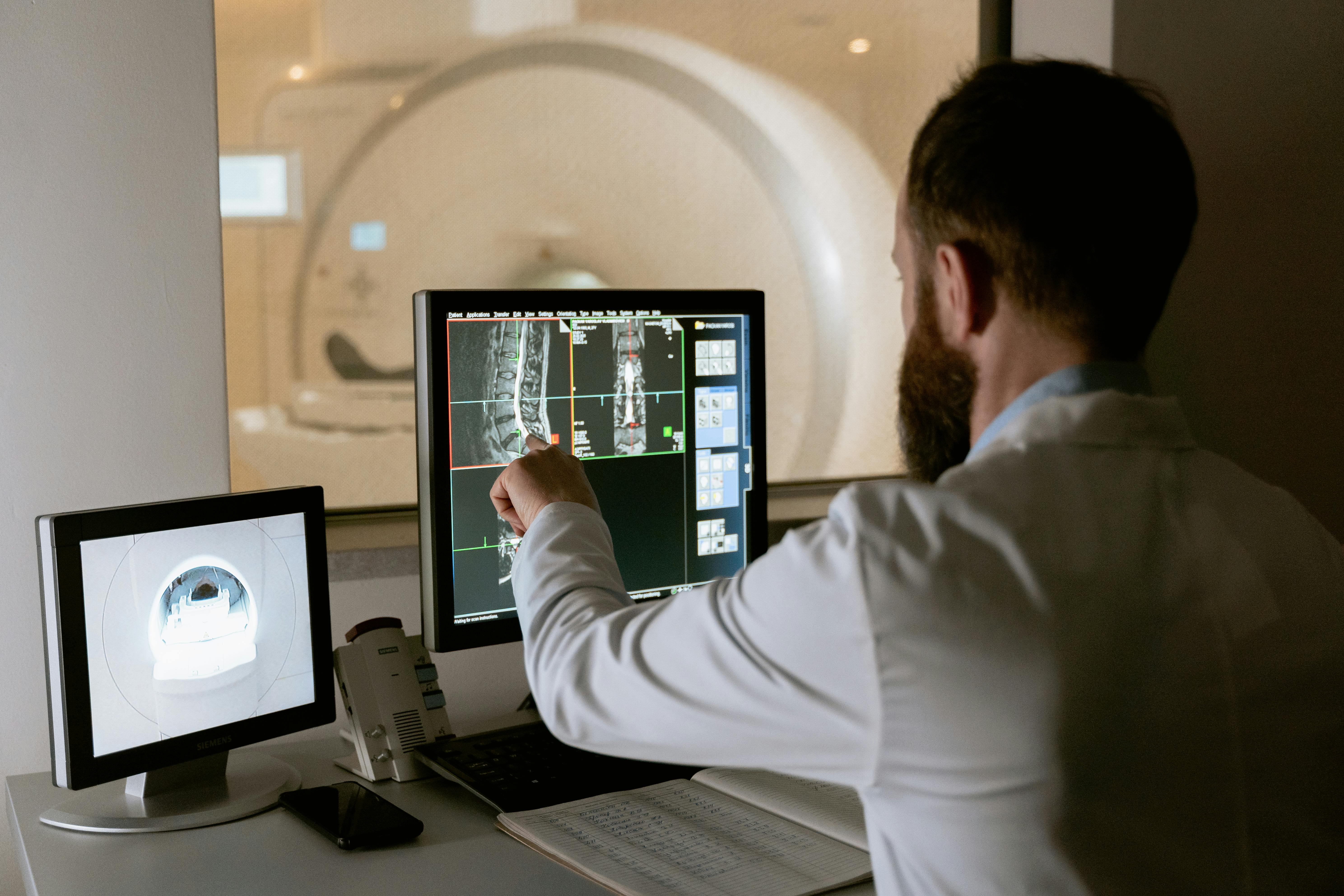 Image of a man monitoring a computer screen that is apparently portraying the results of an MRI. The MRI machine can be seen in the background and on another monitor. 