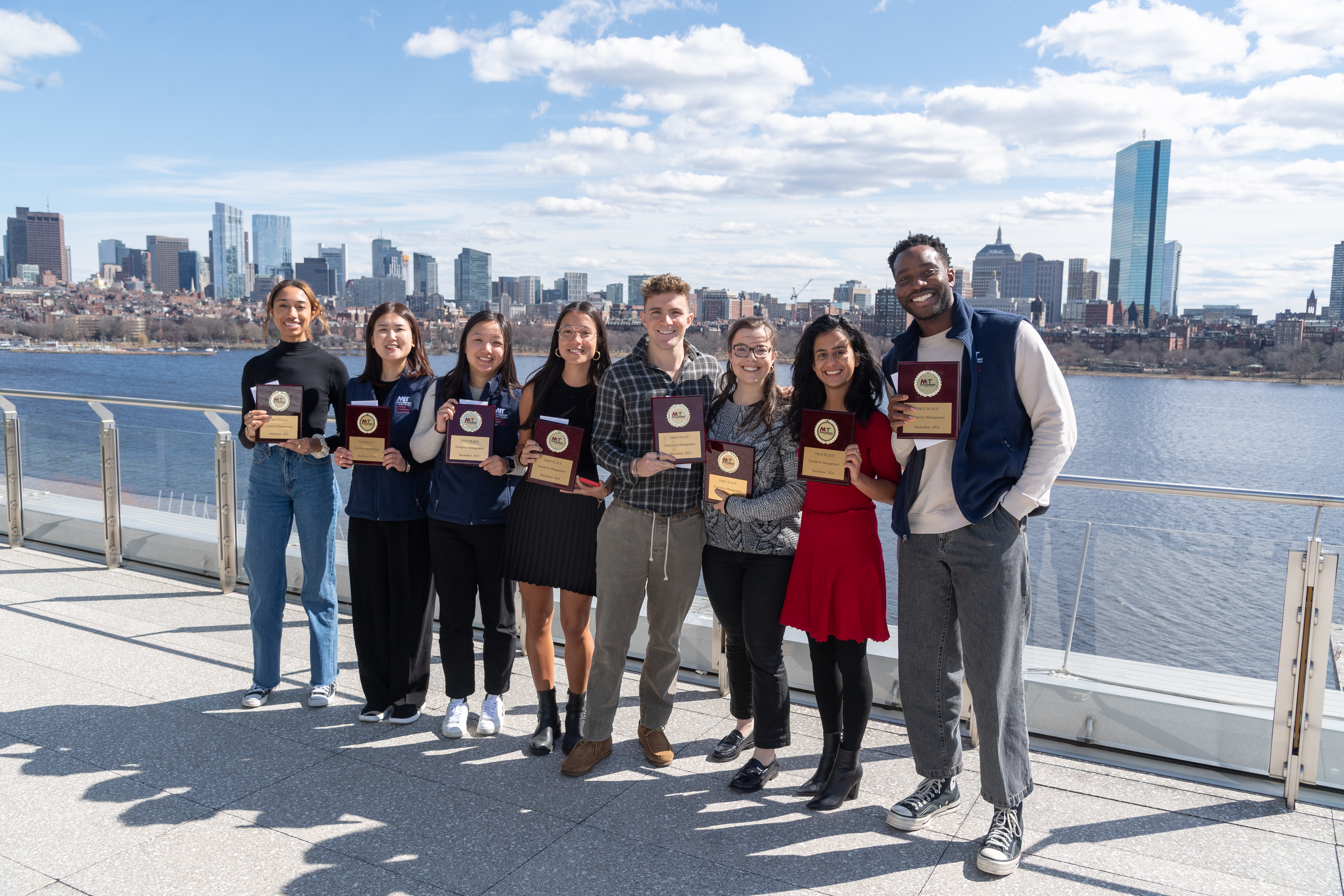 Group photo of the Hackathon 2024 winning team, standing with their plaques with the Charles River in the background.