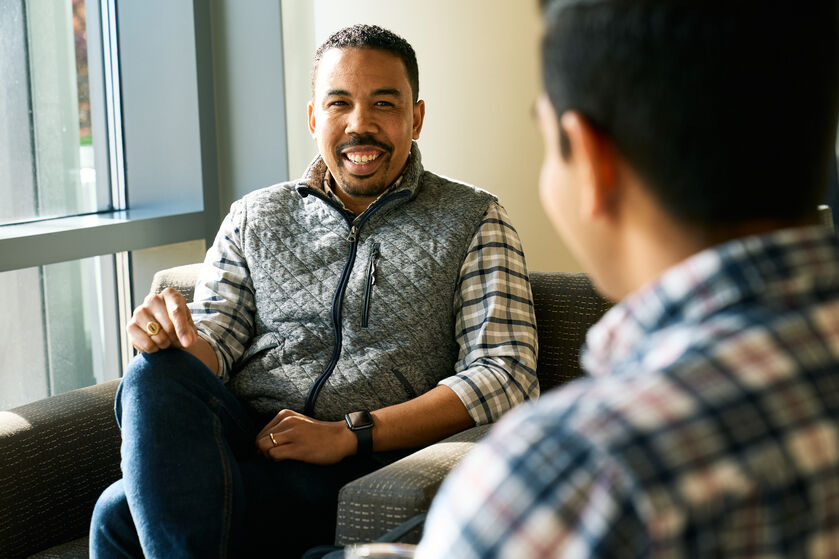 A man with short hair and a vest smiling at his friend.