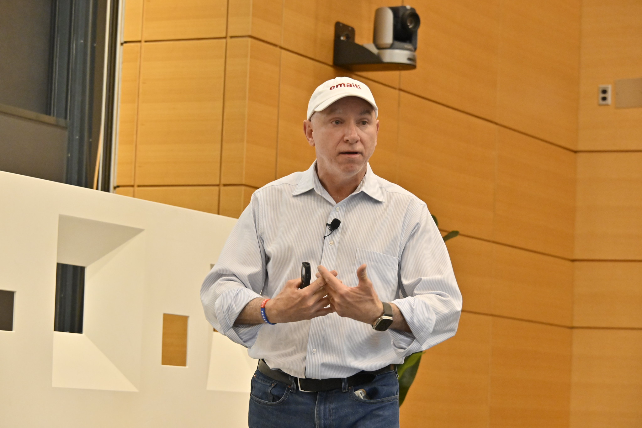 Man in dress shirt, jeans, and hat gestures during presentation.