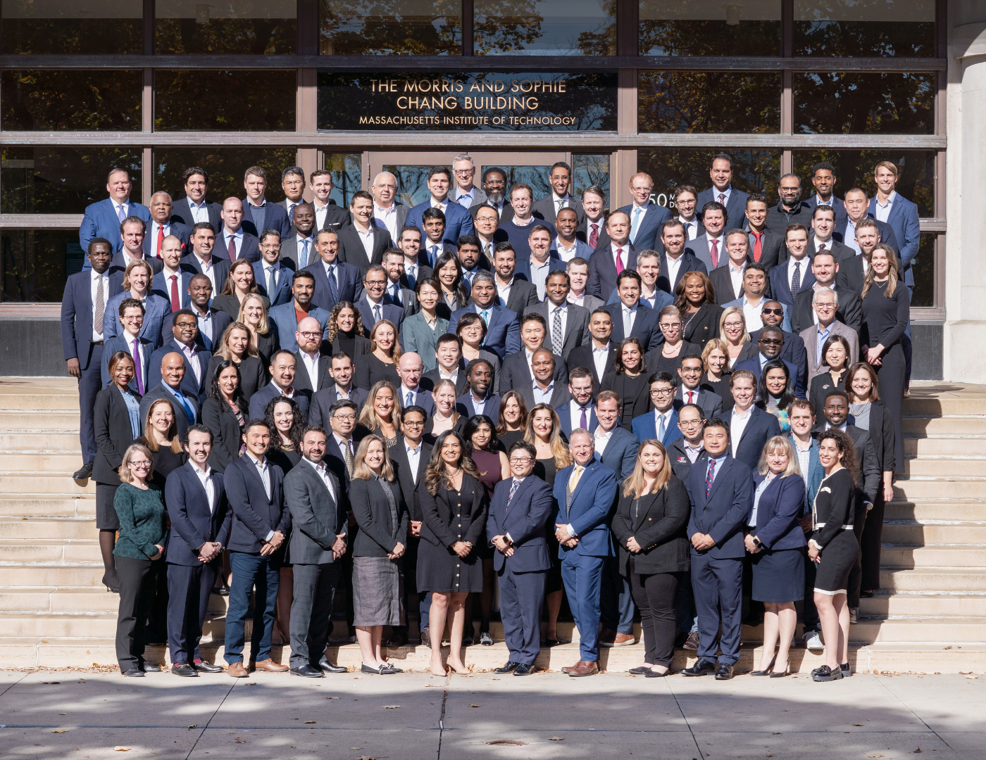 MIT EMBA Class of 2026 standing on the stairs in front of the Memorial Drive Entrance to E52. They are wearing dark business attire.