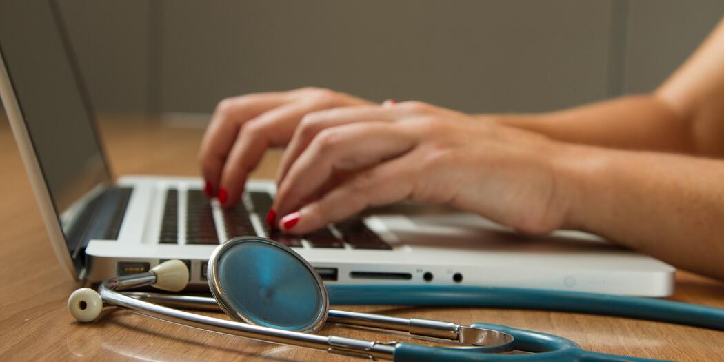 Hands with red nail polish typing on laptop with stethoscope next to it