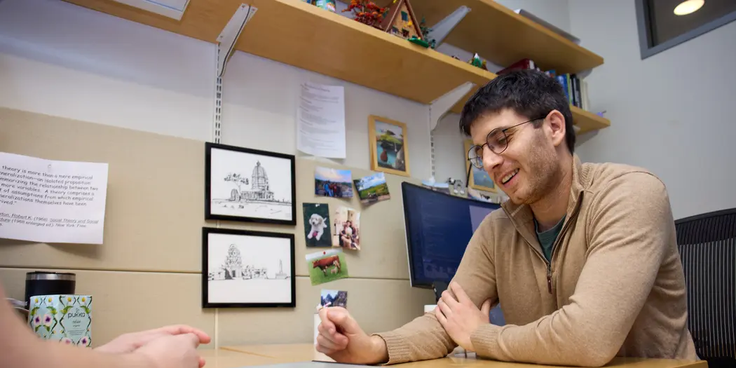 PhD student Ben Manning sits at a desk in the MIT Sloan PhD offices. He looks down at a tablet, talking to someone out of frame.