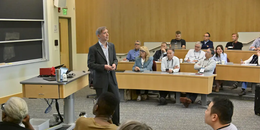 A man in a business casual suit stands before an audience in a classroom