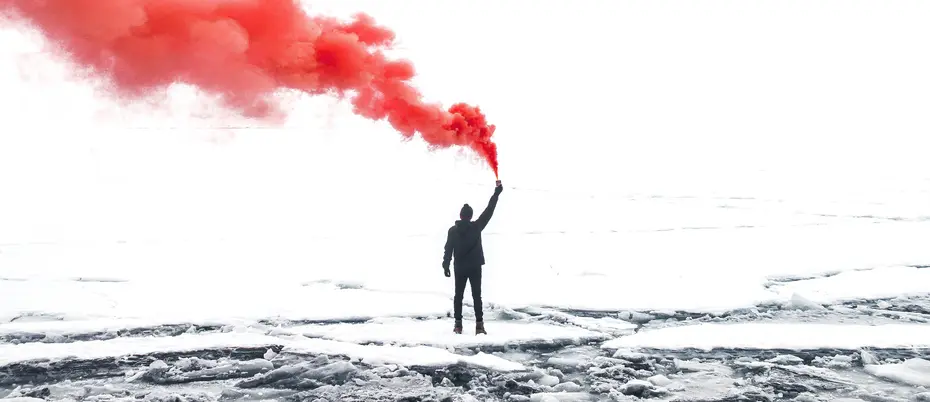 a lone figure stands on a snow-covered rocks, holding a red smoke flare