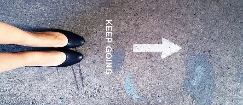 A women in round-toe shoes on a cement floor with the words "Keep Going" and a directional arrow to the right, shot from above.