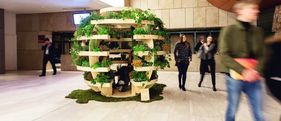 a large wooden sphere with green plants on it sits in a large company lobby with people walking by