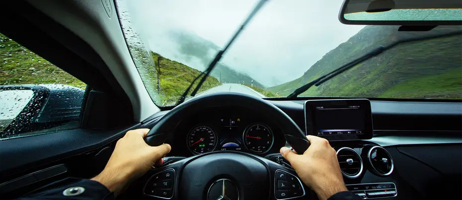 view from the driver's seat of two hands on a steering wheel and a rainy mountain road
