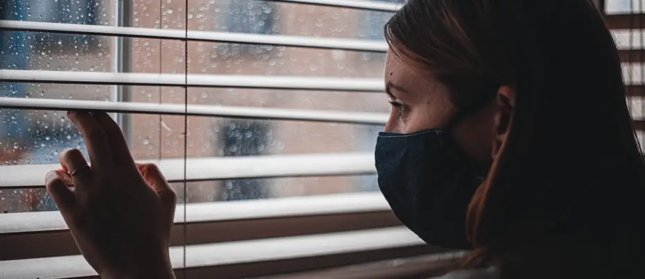 Woman in mask looks out window through blinds.