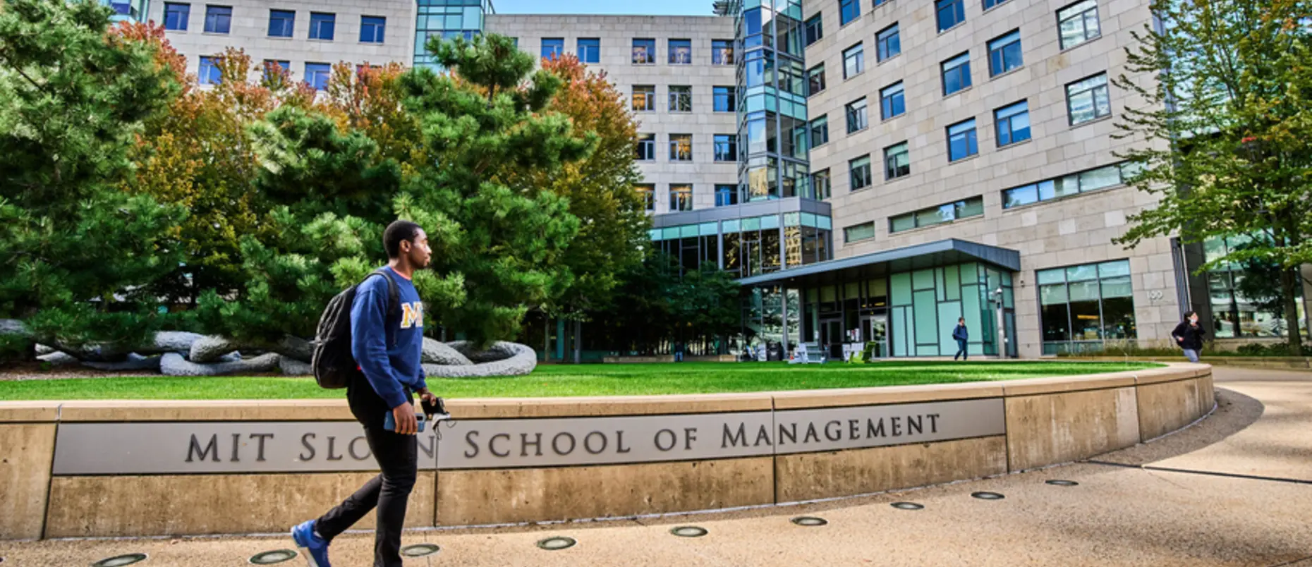 Student walks by the MIT Sloan School of Management sign out front of building E62 on a sunny day
