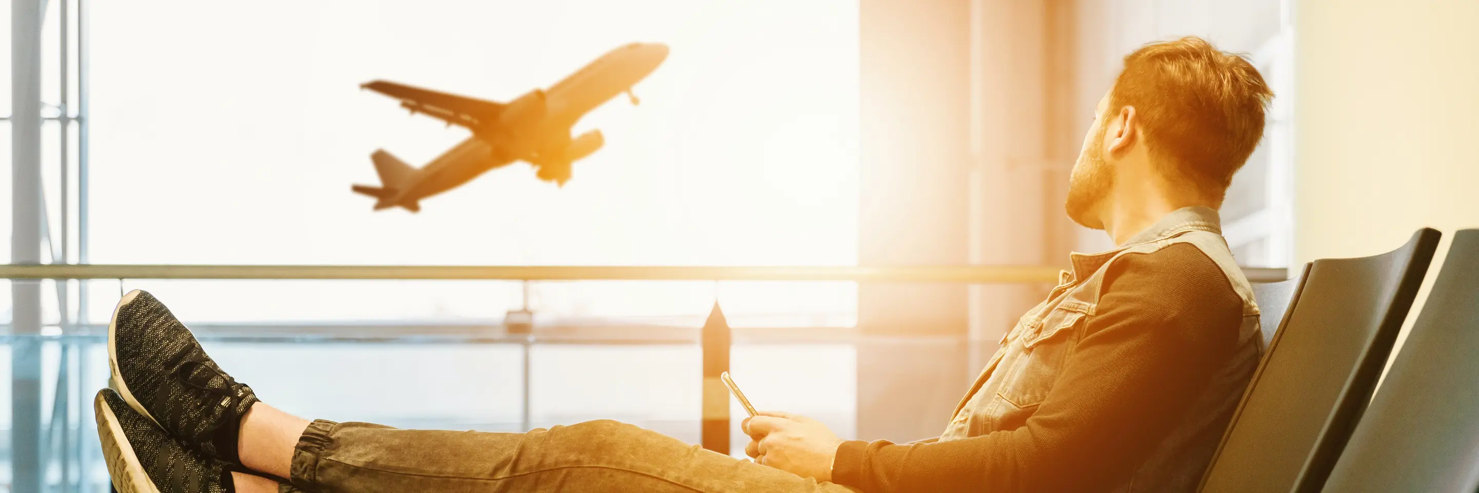 Man sitting in airport waiting area, staring out window at a plane taking off