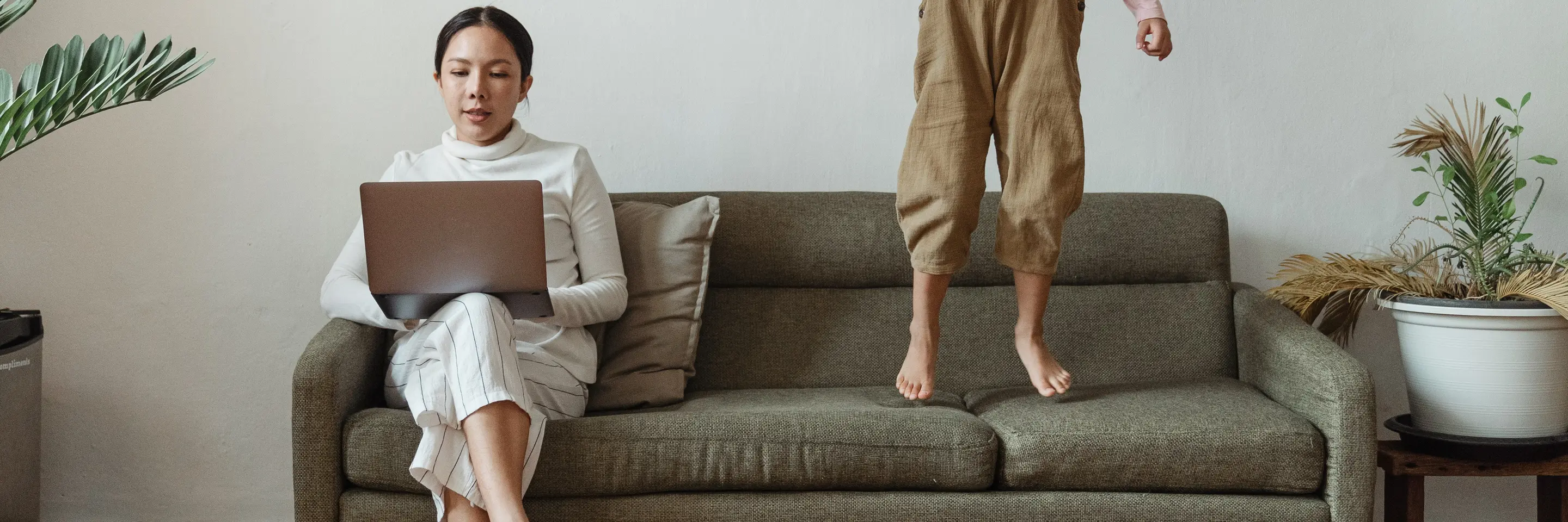 Image of a woman seated on a sofa and working at a computer while a child jumps on the sofa