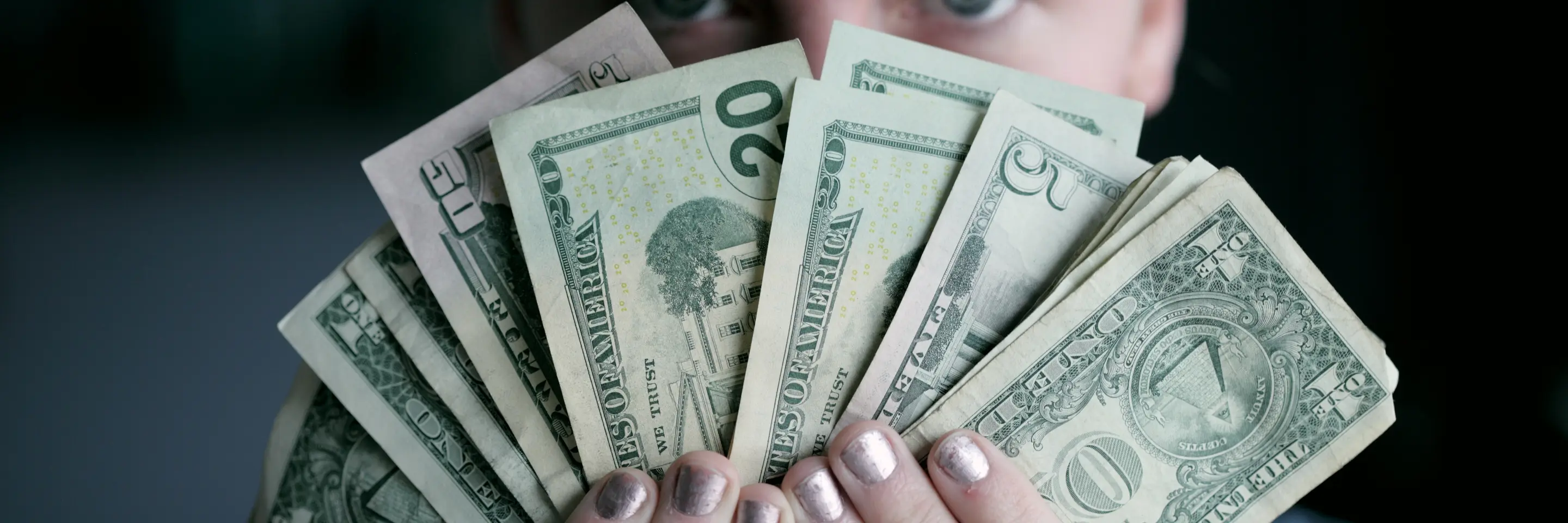 Image of the face of a woman who is holding U.S. currency fanned out in her hands, obscuring the lower half of her face. 