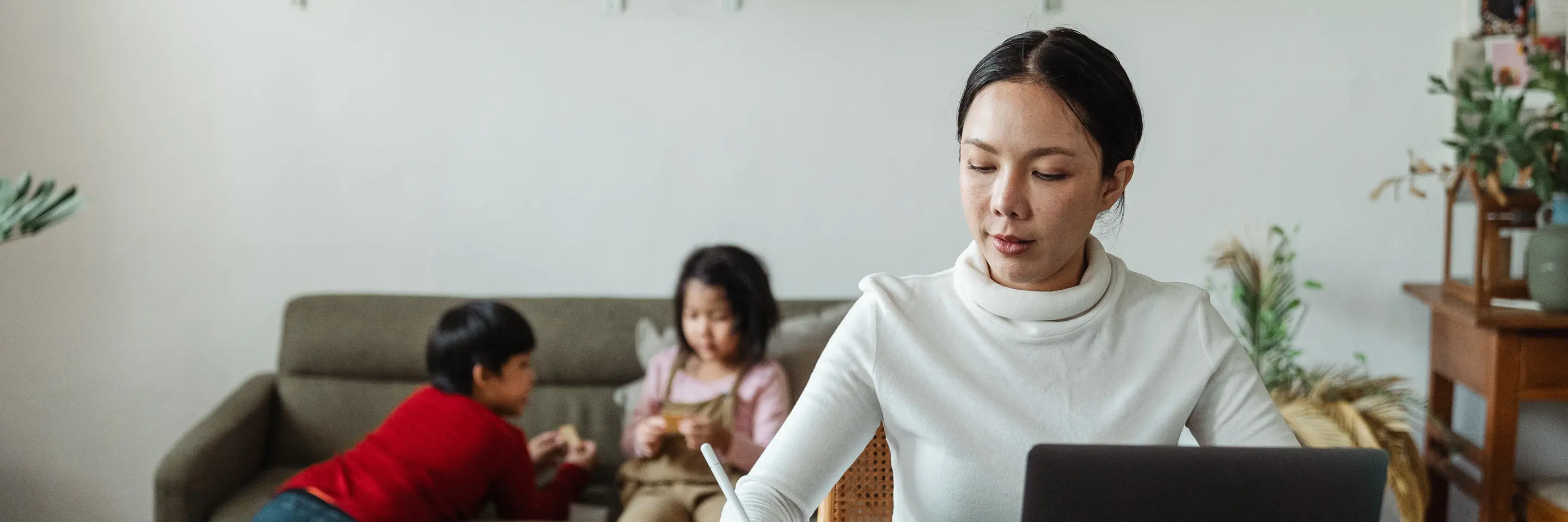 Image of an Asian woman working at home at a computer with two children playing in the background