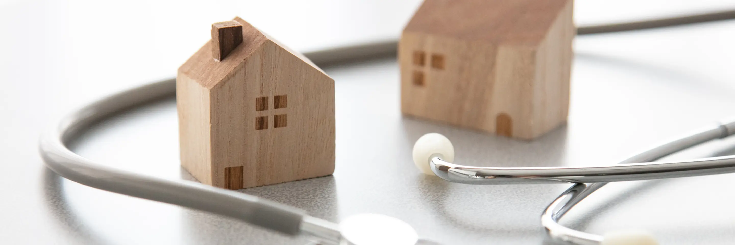 Two mini wooden house figurines with a regular sized stethoscope wrapped around them on smooth white surface