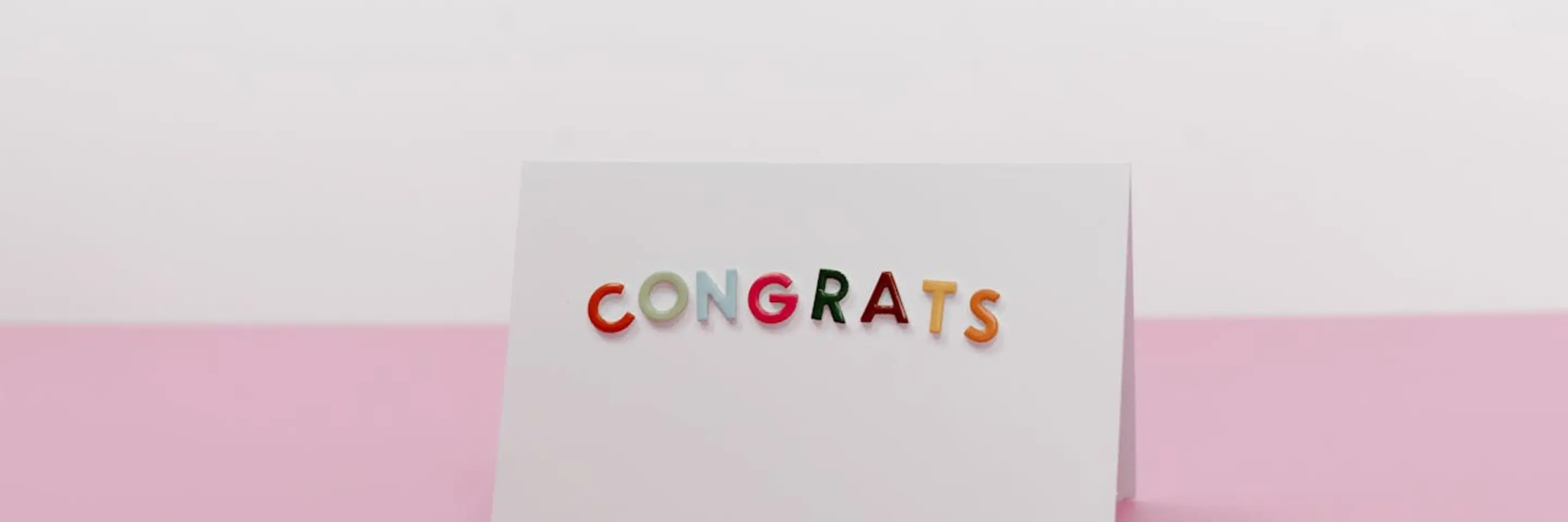 A white card with the word "congrats" on it in colorful letters, against a pink-and-white background