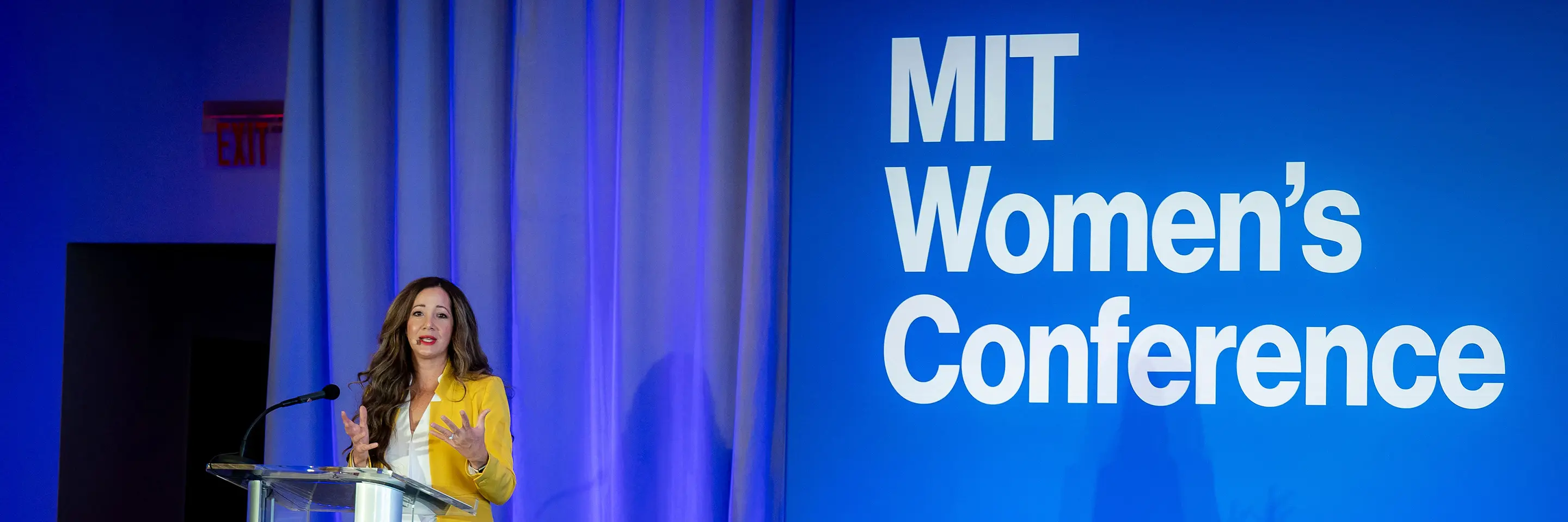 Woman in yellow jack speaks in front of sign for MIT Women’s Conference