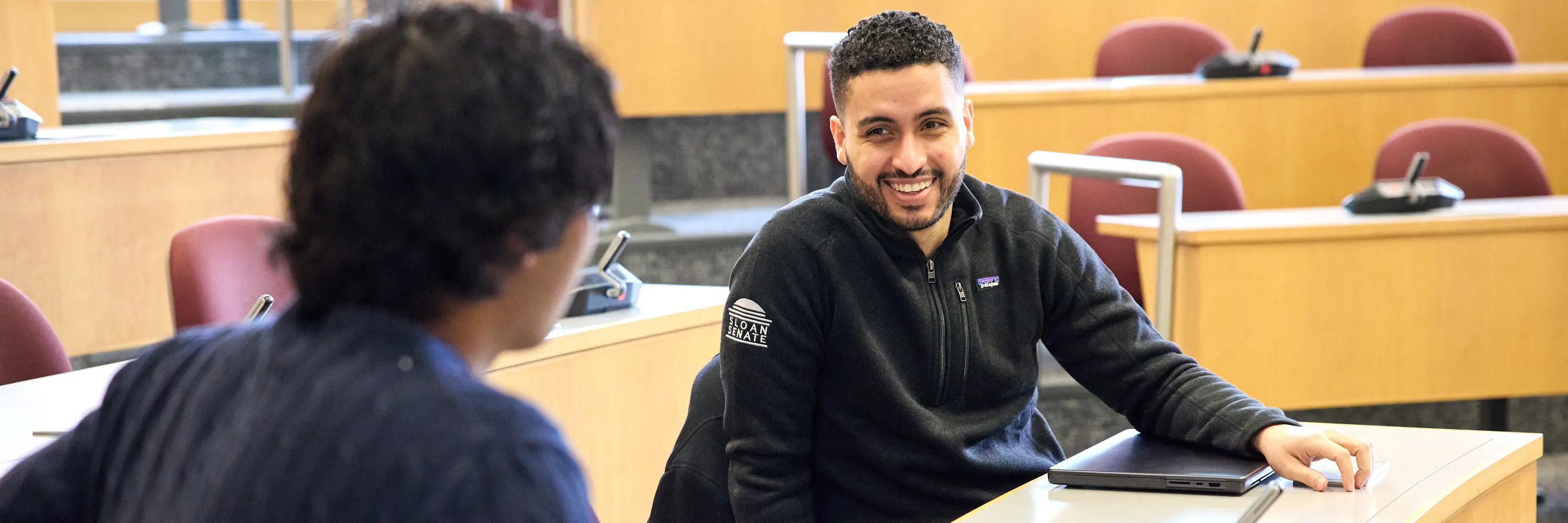 Mike Sanchez sits in an MIT Sloan lecture hall. He smiles at another person, whose back is to the camera