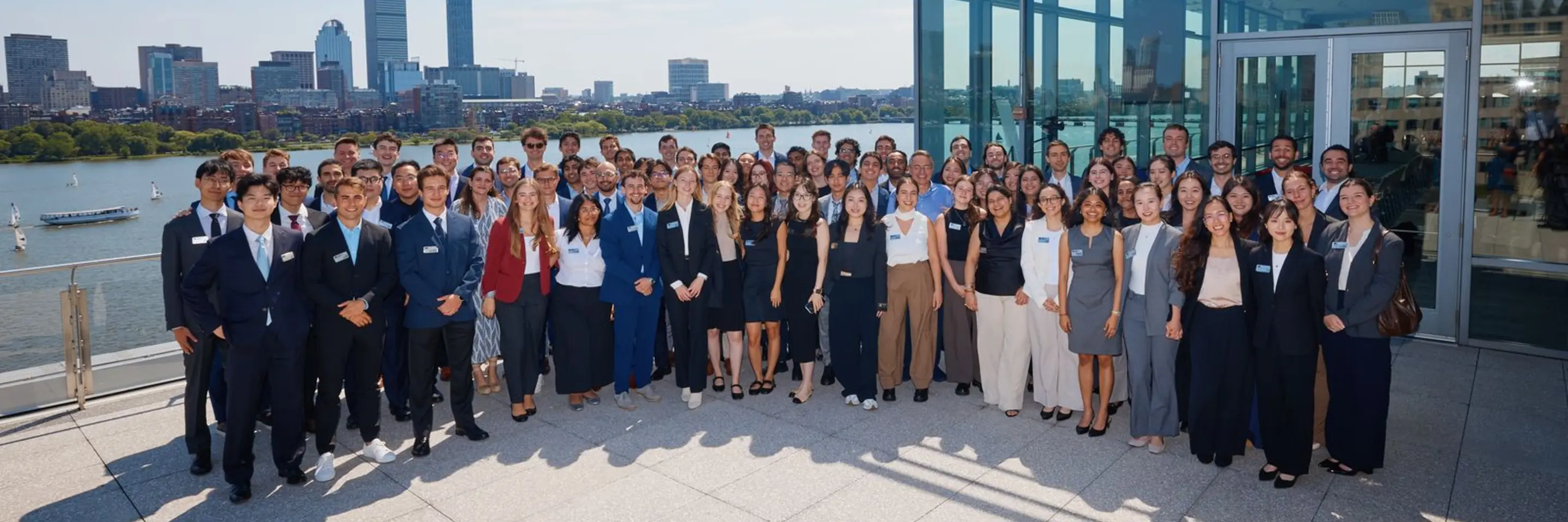 The MBAn Class of 2025 lines up for a photo outside with the Charles River and the Boston skyline in the background