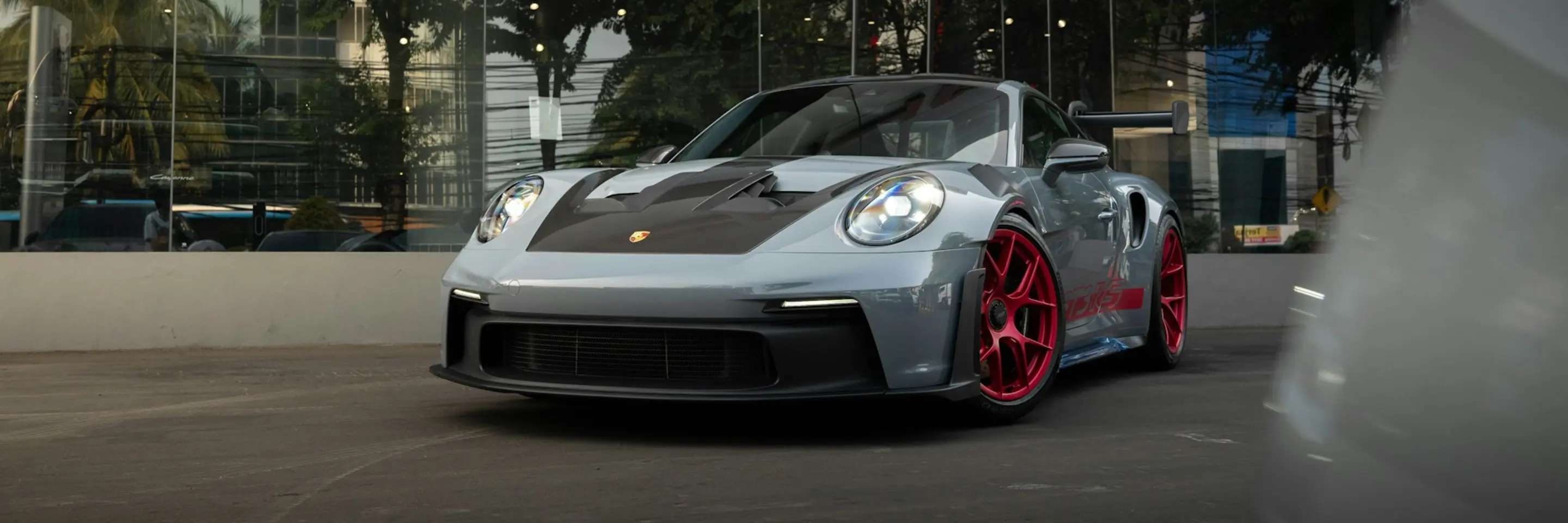 Photo of silver Porsche with a black hood and red rims parked outside of a glass storefront.