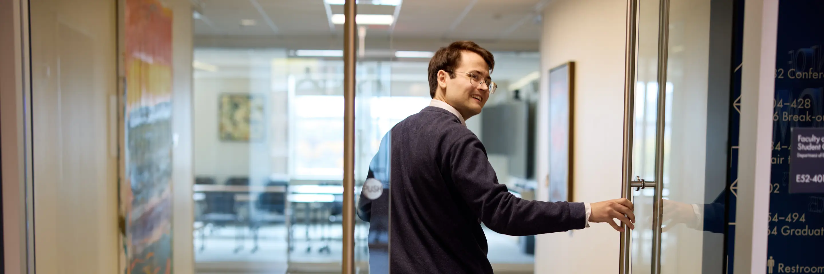 PhD student Alex Busch holds open a door and smiles over his shoulder as he walks through a building at MIT Sloan