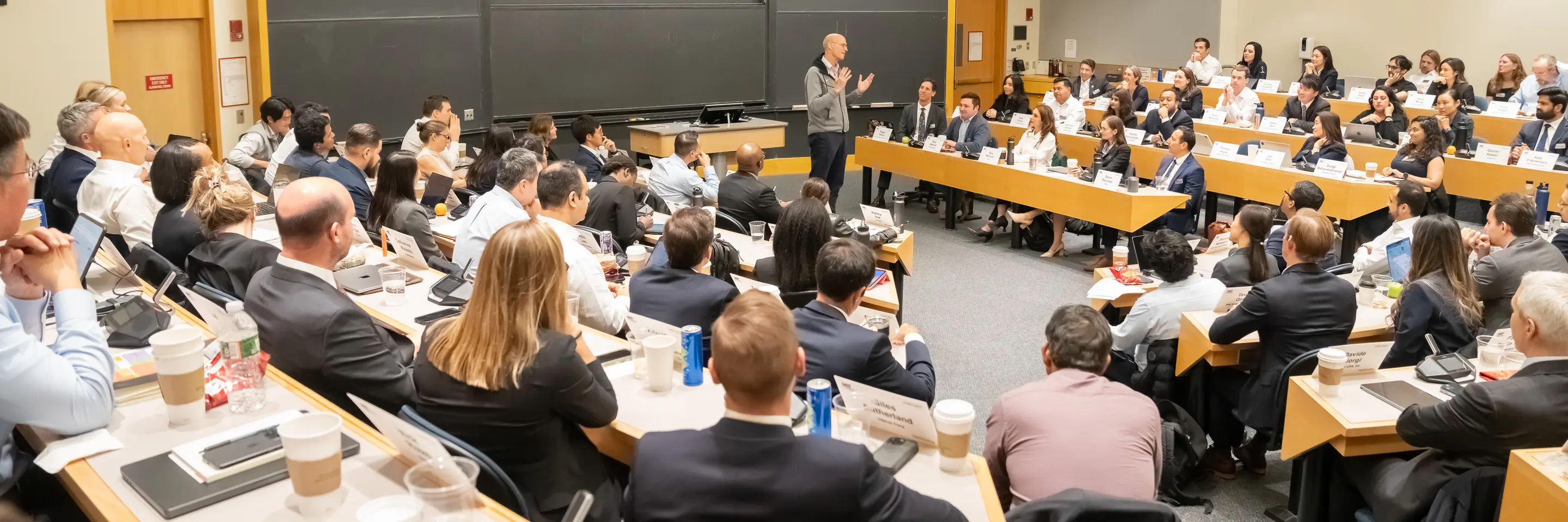 Wide shot of a classroom filled with students facing professor lecturing up front. Behind the professor are empty blackboards.