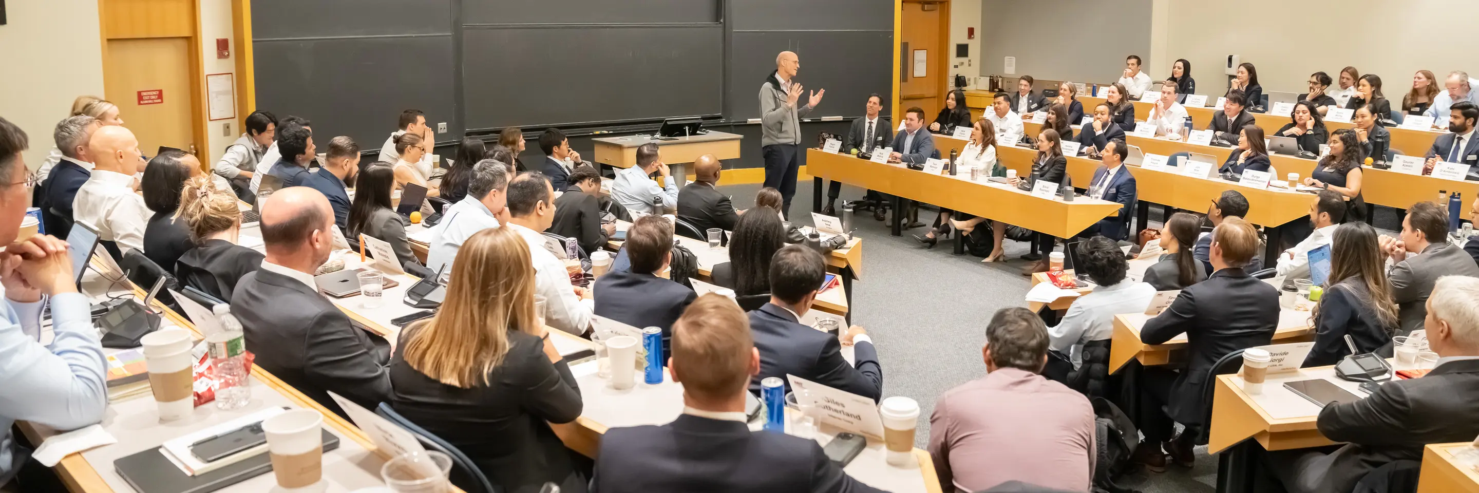 Wide shot of a classroom filled with EMBA students with a lecturer presenting in front of blank black boards