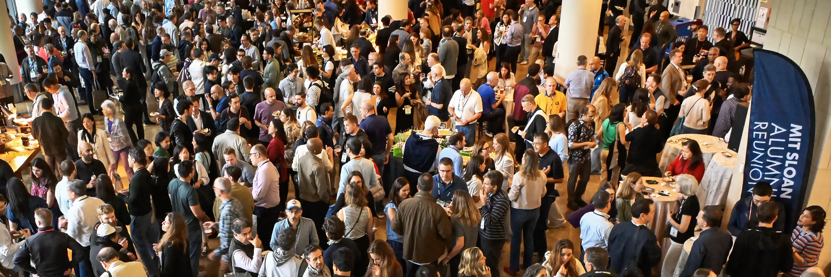 Large crowd gathers in a meeting space nearby banner that reads MIT Sloan Alumni Reunion