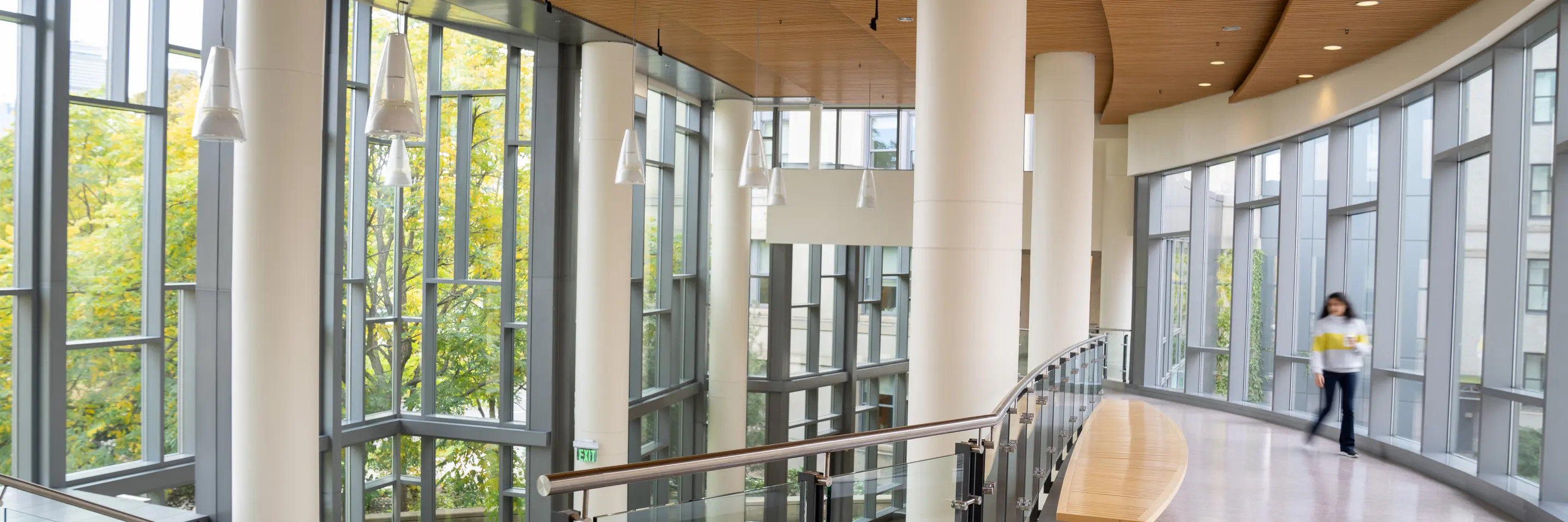 Empty second floor hallway in e62. To the left is a staircase down to the first floor. To the right is the bridge that leads to building E52 if you keep walking straight. There is a woman out of focus walking on the bridge towards the camera all the way to the right of the bridge.