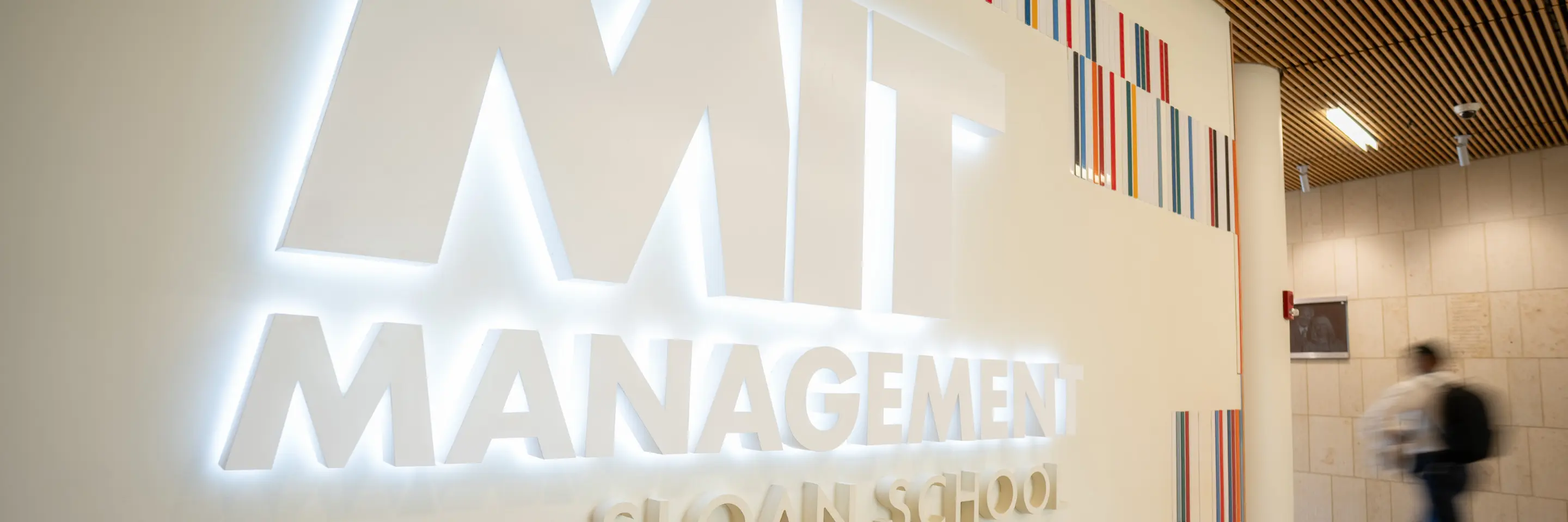 Close-up of the illuminated MIT Sloan School of Management sign and colorful wall accents, with a blurred person walking in the background.