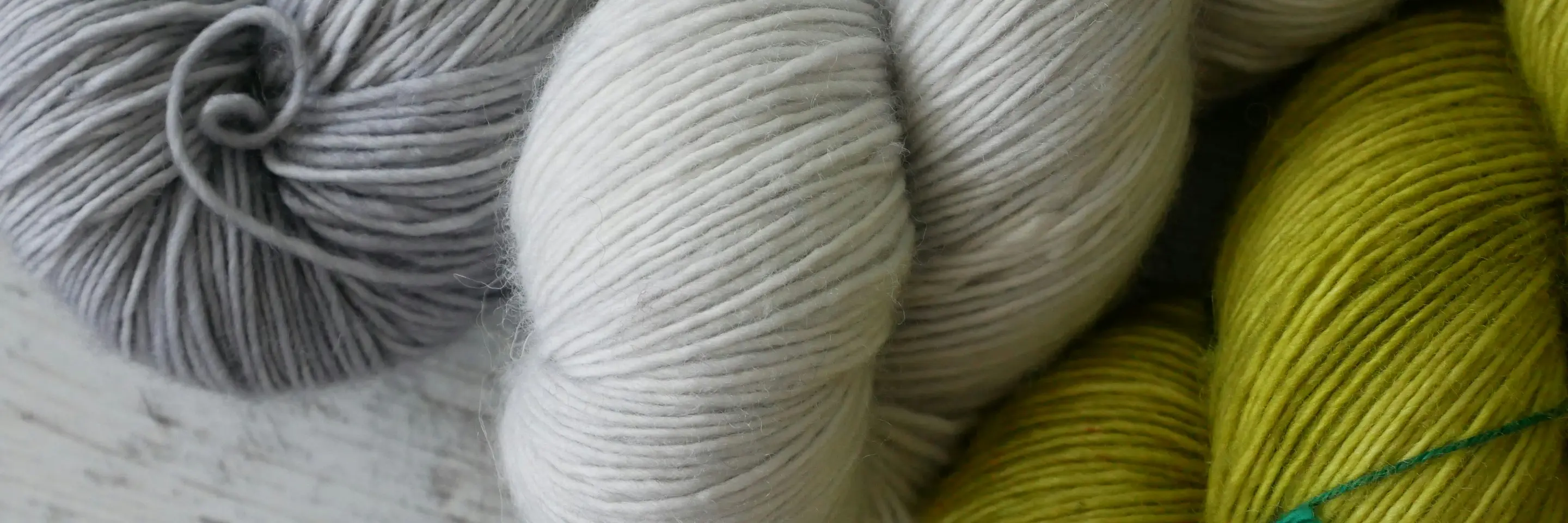 Photo of 3 skeins of yarn: 1 silvery gray, 1 white, and 1 leaf green, sitting on a gray wooden surface