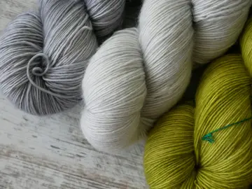 Photo of 3 skeins of yarn: 1 silvery gray, 1 white, and 1 leaf green, sitting on a gray wooden surface